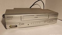 Front view of Magnavox VCR showing front-loading cassette slot, power and function buttons, LED indicator, and input/output ports on bottom left.