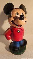 Front view of one Mickey Mouse ceramic piggy bank figurine standing on green base, red shirt with green button, blue pants, black shoes, with signs of light wear on paint.