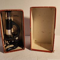 Microscope fitted inside original cardboard box with lid open, showing overall condition and size of the microscope relative to the box.