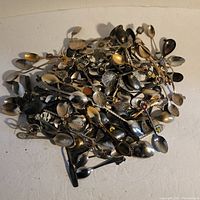Top-down image showing a large assortment of metal spoons piled together. The variety includes spoons with decorative handles with enameled and engraved details.