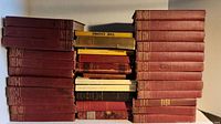Overall view of the stacked vintage books showing many Charles Dickens volumes with red hardcovers and some other books in various colors, dusty with worn edges.