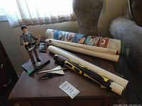 Wide view of the John Lennon figurine, rolled posters, and Concorde model on a wooden table near a window.
