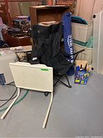 Lot contents overview showing black camping chair, white folding table, blue Handy Bed bag, and box of outdoor lights in basement setting.