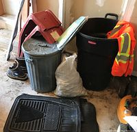 Photo of car ramps, two garbage bins (aluminum and black plastic Rubbermaid), bag of garden rocks, black rubber car mats, men's Kodak steel toe boots, and red under car trolley with orange safety jacket on one bin