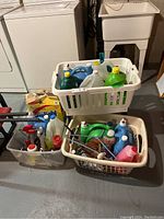 Photo shows three plastic laundry baskets filled with assorted bottles of cleaning supplies and chemicals, including sprays and liquid detergents.