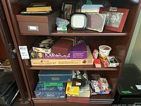 Three shelves showing games and frames with a mix of board game boxes, playing card boxes, and plastic containers. Items arranged with visible labels and tags.