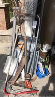 Wide view of garden and snow removal tools including large metal scoop shovel, aluminum shovel, hand saws, hack saws, rake, salt spreader bucket, and men's ice traction slip-ons.