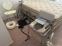 Photo showing white bath chair, gray padded commode seat with armrests, black bed rail, wrist brace, walking canes, padded toilet seat cushion, medical clamp device, all near a mattress on a bed frame
