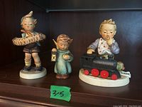 Front view of three Hummel figurines showing a boy with a cob of corn, a girl with a candle lantern, and a boy with a toy train