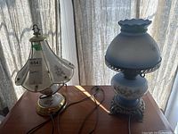 Two table lamps on wood surface near window with sheer curtains, one with cream and floral shade and the other with blue floral shade and ornate metal base.