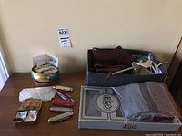 Overall lot view showing leather shoes, scarves on top, leather wallet, tins with stamps and pennies, and pocket knives on table.