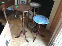 Three plant stands shown together. From left to right: wooden stand with round top, cane woven top stand with brass accented base, and white marble top stand with red wooden base.