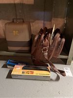 Hard plastic beige and brown case with yellow 'K' emblem, leather camera bag with multiple zippered compartments, and two boxed Sennheiser accessory items on concrete floor shelf.