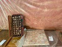 Full view of stone chessboard and chess pieces in original cardboard packaging, on a wooden surface near a pink-tied backdrop.