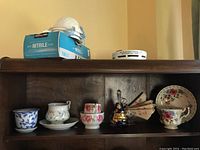 Photo showing a wooden shelf holding various tea cups including Royal Albert and Bell China sets, plus an art glass basket and some other ceramic cups.