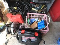 Overall shot showing electric Skilsaw with circular blade and guard, pink basket with soldering kit and cords, black power cords, screwdrivers, and Black & Decker zip saw case