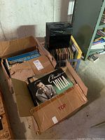 View of three containers with vinyl records including a box with Claudio Villa album on top, a black milk crate filled with records, and a separate cardboard box. Setting is a basement with concrete floor and bare walls.