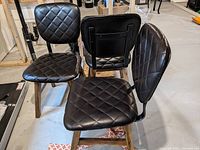 Photo of three black leather-like chairs with wooden legs and metal back supports arranged in a room with concrete floor.