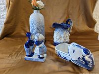 Photo showing four blue and white houseware items: Holland kiss figurine, tall vase with bubble pattern, candy or soap dish with lid showing windmill design, and ornate Staffordshire ironstone jug with embossed details.