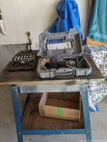 Dremel Trio rotary tool and mini ratchet set placed on a wooden table. Hard plastic case containing the rotary tool with various accessories visible. Black molded tray holding mini ratchet and sockets placed beside the Dremel case.