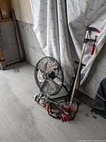 Floor fan, small sander, five small clamps, one large clamp, and a hand pump visible on concrete floor against a wall with a draped cloth.
