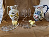 Photo showing two ceramic pitchers with floral designs, six clear champagne flutes, two clear glass plates each with yellow chick-shaped egg cups.