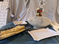 Photo showing folded yellow cloth napkins in a dark metal tray, white ceramic rectangular serving dishes, and a decorative vase with floral design and gold neck holding artificial flowers.