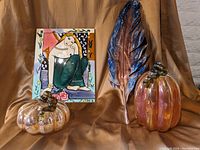 Photo shows ornamental tin feather, two colored glass pumpkins, and a colorful artistic cutting board against brown background.