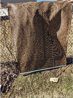 Photo of brown outdoor woven blind with weighted bottom bar and pull handle attached by cord, displayed outdoor against chain link fence with natural daylight.