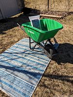 Green Praktika wheelbarrow standing on a patterned blue and white outdoor carpet, outside in yard under sunlight. Packaging for sun deck sail seen inside wheelbarrow.