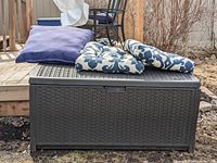 Black plastic waterproof bench with woven rattan texture outdoors. Four cushions on top: two solid blue and two white with blue floral prints.