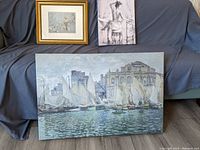 Three picture prints displayed on a sofa, including a Claude Monet seascape with sailboats and a building, a figurative sketch/painting of a person sitting on a stool, and a nature-themed print by Robert Bateman in a gold fleck frame.
