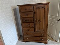 Photo shows a pine dresser against a white textured wall and a white door. The dresser features six drawers on the left and one cabinet door on the right with round wooden knobs. It has a natural pine wood finish with visible knots and wood grain, standing on a block base.
