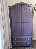 Full frontal image of the purple painted pine wardrobe featuring two doors and two bottom drawers with white knobs, arched top and paneled fronts.