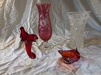 Four glass items arranged on light fabric backdrop: cranberry floral etched vase, heavy crystal cut vase, small red curved glass piece, and red/orange glass basket with handle.