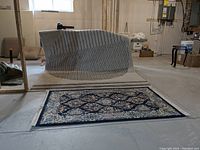 Photo showing two rugs on a concrete floor in a basement setting. The large rug is draped over a stack with black and cream stripes. The smaller rug lies flat with a blue border and floral design.