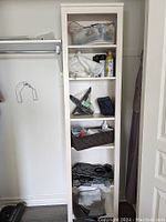 White wooden storage shelf tower standing in a closet, holding miscellaneous household items for display only, contents not included in sale.