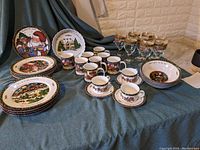 Wide view of the Christmas-themed porcelain dinnerware and glassware arranged in groups showing mugs, plates, bowls, cups with saucers, and wine glasses on a blue fabric surface against a brick wall backdrop.