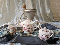 Porcelain tea set including teapot, sugar bowl, and creamer on a blue and black striped fabric table runner with tassels.