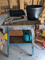 Utility table with metal frame and wooden top, large black plastic plant pot, watering can, bar clamps, spring clamp pliers, and sharpening tool in a workshop setting