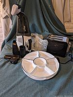 Photo showing Curtis Stone toaster, Shark handheld vacuum with attachments, two white segmented plastic party plates, and vacuum accessories on green fabric background.