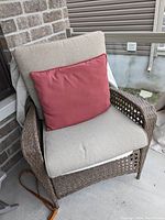 Brown wicker outdoor chair with beige seat and back cushions and a red accent cushion on a concrete patio by a brick wall.