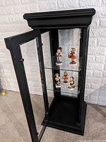 Black painted wood display cabinet with glass door open showing two glass shelves with five Hummel figurines inside