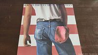 Front cover of Bruce Springsteen Born in the U.S.A. vinyl LP showing a man in jeans with a red cap against a red and white striped background.