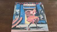Front cover of the vinyl album "She's So Unusual" featuring Cyndi Lauper in a pink dress roller-skating in front of a blue building.