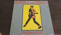 Front cover of Elvis Costello album titled 'My Aim Is True' showing image of Elvis Costello in a salmon jacket on checkerboard background.