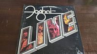Front cover of the vinyl album 'Live' by Foghat, showing band member photos within the title and an autograph in black marker.