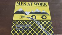 Front cover of business as usual vinyl album by Men At Work with yellow and black artistic design featuring mountains and guitar.