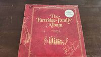 Front of the red vinyl album cover showing the title 'The Partridge Family Album' with musical notes, gold lettering, signature from Shirley Jones and David Cassidy, and visible wear and damage around edges.