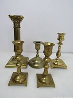 Photo showing front view of five solid brass candle holders of various heights and designs placed on white surface.